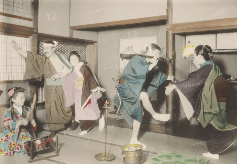 Three geishas and two men dance and make music in a teahouse in Japan, page, photomechanical print, inscription: 'This represents the final scene, after which the entertainers are paid and dismissed, and the guests take themselves home, probably to receive a scolding for being out so late.', maker: Kozaburo Tamamura, Japan, c. 1896 - c. 1906, paper, collotype, light print, hand colored, height 105 mm x width 151 mm, performance, tea garden, garden restaurant, A colorized photograph of a group of people in traditional clothing inside a room with sliding panels and wooden beams, showing a man with a headband and loose robe gesturing with one arm, a seated girl in a patterned kimono near a low brazier, a woman in a light-colored kimono kneeling and smiling, a person in a blue robe balancing on one leg with hands raised, and another woman holding a folded fan while seated, with cushions and small decorative objects on the floor and a hanging scroll on the wall.