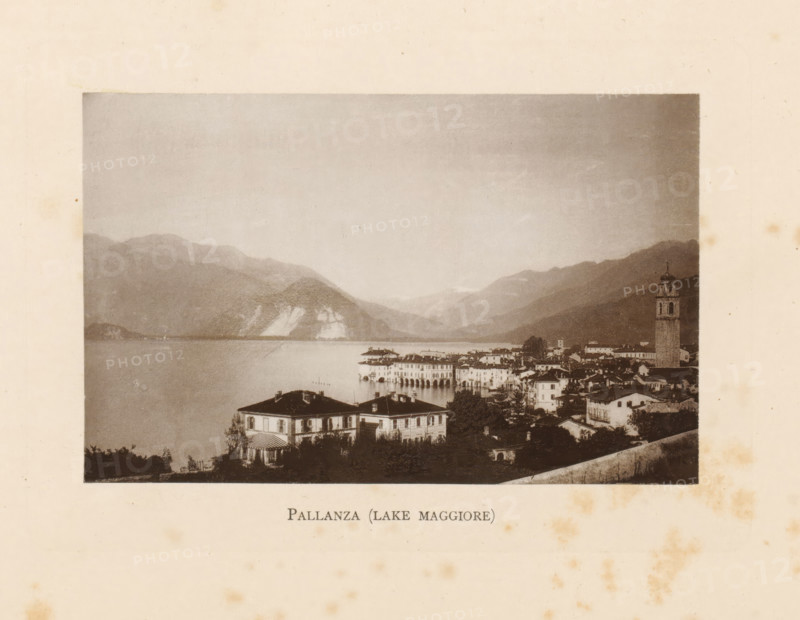 View of Pallanza on Lake Maggiore in Italy, Pallanza (Lake Maggiore), page, photomechanical print, Italy, c. 1874 - in or before 1884, paper, collotype, photoprint, height 86 mm x width 136 mm, lake, prospect of village, silhouette of village, mountains, Piemonte, Verbania, A sepia-toned photograph framed on aged paper shows a lakeside town with clustered buildings and a prominent bell tower rising above the rooftops, a waterfront with arched colonnades extending into the water, and villas with shuttered windows and striped awnings in the foreground; calm water fills the middle ground and reflects the shoreline, while layered hills and mountains fade into the sky in the background.