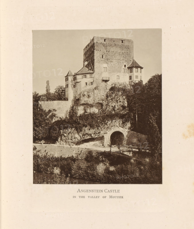 View of Schloss Angenstein, Angenstein Castle in the Valley of Moutier, page, photomechanical print, Basel-Landschaft, c. 1874 - in or before 1884, paper, collotype, photoprint, height 120 mm x width 100 mm, castle, Schloss Angenstein, A monochrome photograph of a large stone castle perched on a rocky hill, showing a tall rectangular keep with small windows and rough masonry, flanked by smaller turrets with conical roofs and multiple windowed facades; vegetation and trees grow around the rocky base, and a rounded arched opening is visible at the rock face leading into a tunnel or passage; a low stone wall and a path run along the hillside below the castle, and the scene is framed by a wide blank border.