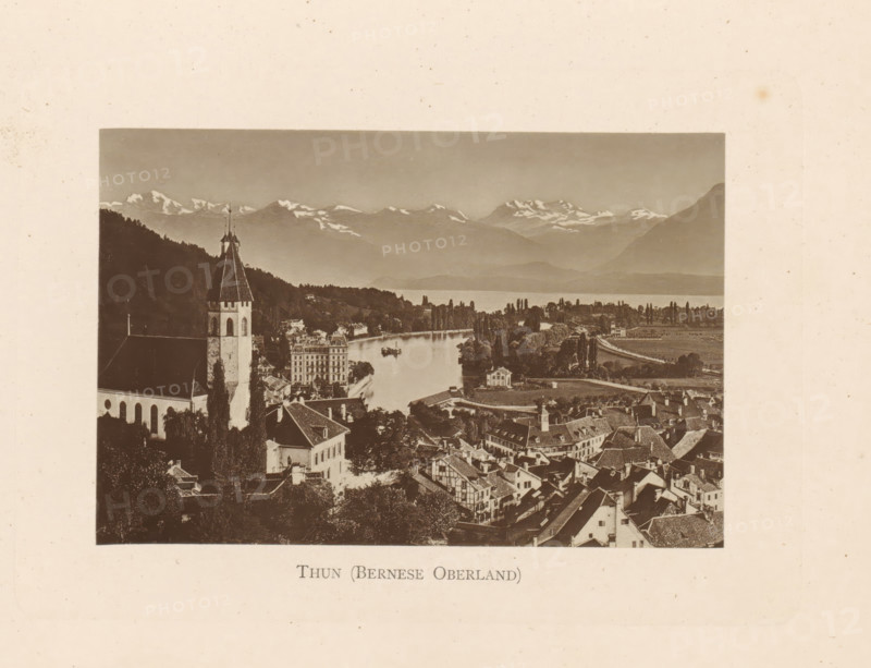 Thun (Bernese Oberland), View of Thun in Switzerland, page, photomechanical print, Switzerland, c. 1874 - in or before 1884, paper, collotype, photoprint, height 85 mm x width 128 mm, city-view in general, 'veduta', mountain-range, snow peaks, A sepia photograph showing a lakeside town with clustered rooftops and narrow streets in the foreground, a tall church tower with a steep pointed roof rising among trees to the left, a river or inlet curving through the middle of the scene with boats on the water, rows of trees along the shoreline and open fields beyond, and distant snow-capped mountains under a clear sky.