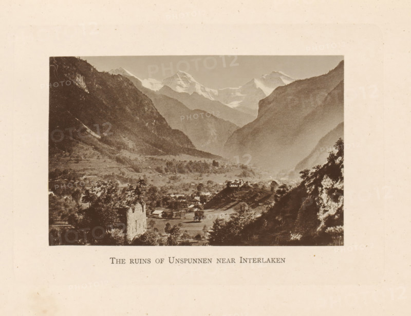 The ruins of Unspunnen near Interlaken, View of the ruins of Burg Unspunnen near Interlaken, page, photomechanical print, Switzerland, c. 1874 - in or before 1884, paper, collotype, photoprint, height 82 mm x width 127 mm, mountains, ruin of a dwelling, house, castle, A sepia-toned landscape photograph framed by a wide margin shows a valley between steep, dark mountain slopes with snow-capped peaks visible in the distance. In the foreground there are scattered trees, open fields, and several small buildings, including a prominent ruined stone structure on a rise and a house perched on a hill. Layers of ridges recede toward the distant glaciers and peaks, and the scene has a soft, hazy atmospheric gradation from the foreground to the distant mountains.