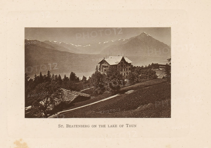 St. Beatenberg on the lake of Thun, View of a house in Beatenberg on Lake Thun, page, photomechanical print, Beatenberg, c. 1874 - in or before 1884, paper, collotype, photogravure, height 80 mm x width 128 mm, lake, mountains, Lake Thun, A sepia-toned landscape photograph bordered by a pale background shows a large multi-story chalet-style building on a hillside surrounded by trees and smaller wooden structures, with a steep grassy slope in the foreground and a winding path leading toward the building; behind it a range of mountains rises with snow patches on ridgelines and a prominent peak to the right, and a band of darker forested hills fills the midground beneath a pale sky.