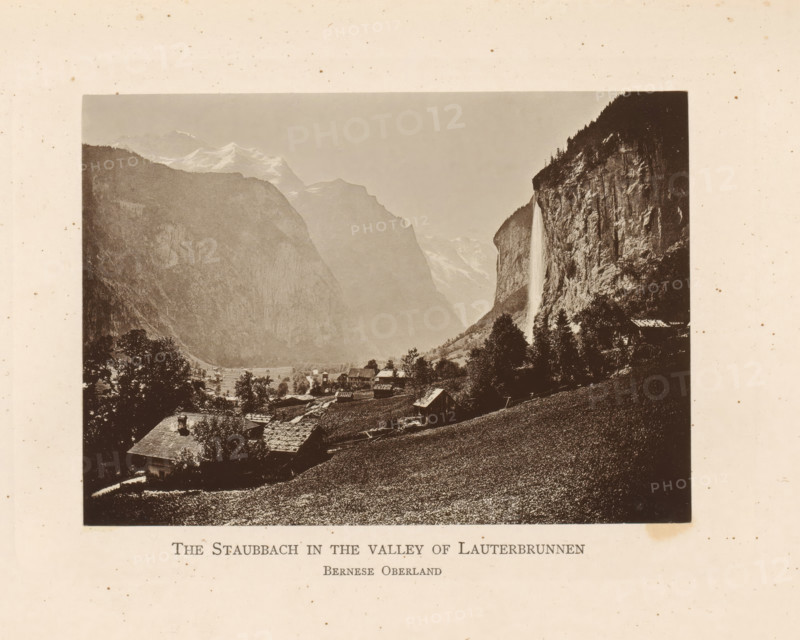 View of the Lauterbrunnen Valley with the Staubbach Waterfall, The Staubbach in the Valley of Lauterbrunnen Bernese Oberland, page, photomechanical print, Bern (canton), c. 1874 - in or before 1884, paper, collotype, photoprint, height 93 mm x width 131 mm, waterfall, dale, valley, Lauterbrunnen, Staubbachwaterval, A sepia-toned photograph shows a valley framed by steep rocky cliffs and distant snow-capped peaks under a pale sky, with a tall narrow waterfall cascading down a cliff face on one side; below, clustered rustic buildings with pitched roofs sit among trees and sloping grassy fields, and a foreground hillside covered in textured vegetation slopes toward the village.