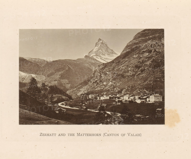View of Zermatt and the Matterhorn, Zermatt and the Matterhorn (Canton of Valais), page, photomechanical print, Valais (canton), c. 1874 - in or before 1884, paper, collotype, photoprint, height 85 mm x width 130 mm, mountains, Zermatt, Matterhorn, A sepia-toned landscape photograph framed by a wide pale border shows a mountain peak with a jagged, snow-covered summit rising above steep rocky slopes; below the peak a valley contains clustered buildings including a church with a steeple and several multi-story structures, a winding river or stream flowing through fields and past small trees, and wooded hillsides and rocky outcrops on either side.