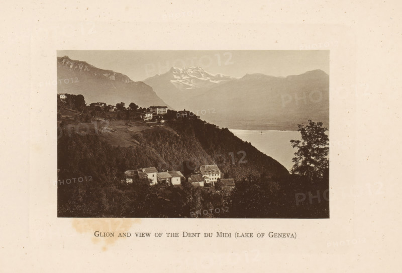 Glion and view of the Dents du Midi (lake of Geneva), View of Glion on Lake Geneva and behind it the Dents du Midi, page, photomechanical print, Montreux, c. 1874 - in or before 1884, paper, collotype, photoprint, height 83 mm x width 134 mm, lake, prospect of city, town panorama, silhouette of city, mountains, Glion, A sepia-toned photograph shows a lakeside mountain scene with a foreground of clustered houses on a forested hillside, a steep dark ridge descending toward a body of water, a tree silhouette at the right edge, distant misty mountains across the lake with a snow-capped peak visible toward the center, and a pale sky above.