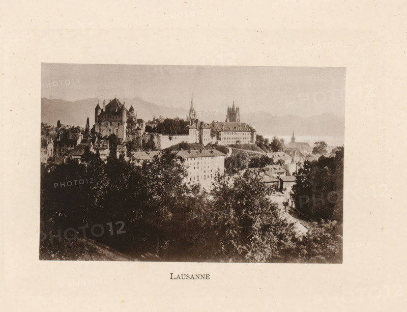 Lausanne, View of Lausanne in Switzerland, page, photomechanical print, Vaud (canton), c. 1874 - in or before 1884, paper, collotype, photoprint, height 83 mm x width 128 mm, city-view in general, 'veduta', A sepia-toned photograph shows a hillside town with clustered buildings, rooftops, and trees in the foreground and midground, a large castle-like structure with turrets and a high wall to the left, a cathedral with a tall spire and another church with a squared tower toward the center, rolling hills or mountains in the distant background, and a large dark treeline occupying the lower portion of the image.
