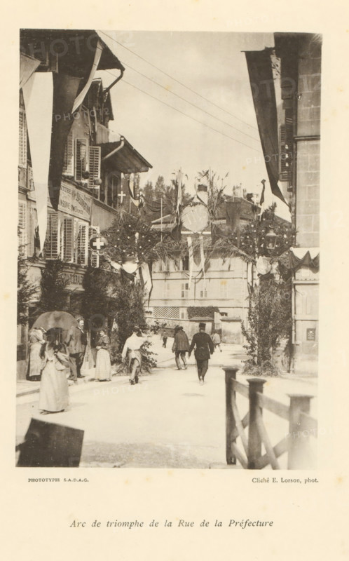 Arc de triomphe de la Rue de la Préfecture, View of a street with an arch of honor in Fribourg, page, photomechanical print, maker: Ernest Lorson, Fribourg, maker: SADAG, 1897, paper, collotype, photoprint, height 162 mm x width 111 mm, street, triumphal arch, Friborg, A sepia-toned street scene framed by buildings on both sides shows an archway decorated with foliage and draped fabric spanning the road, with hanging round ornaments and flags. People in period clothing walk and stand beneath and near the arch, including women in long dresses and a man wearing a uniform with a hat. Shuttered windows and awnings are visible on the façades, and lamp fixtures and potted greenery adorn the street. A wooden railing runs along the right foreground, and a multi-story building with shuttered windows fills the background behind the arch.