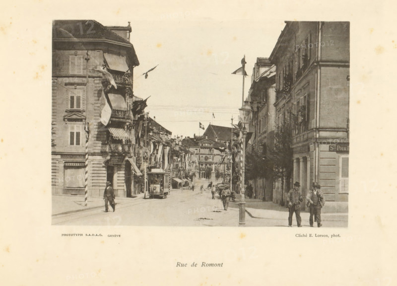 Rue de Romont, View of Rue de Romont in Fribourg, page, photomechanical print, photographer: Ernest Lorson, Fribourg, maker: SADAG, Genève, 1897, paper, collotype, photoprint, height 113 mm x width 162 mm, tram, street, advertising, A monochrome street scene framed by tall buildings on both sides, with awnings and balconies projecting over the sidewalk and flags hanging from several facades, a tram car on tracks near the foreground, pedestrians walking along the street and standing on the sidewalks, a lamppost or pole rising near the center of the image, architectural details such as shuttered windows and decorative stonework on the buildings, distant buildings visible down the street and a sky above.