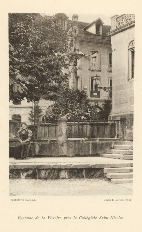 View of a fountain in Fribourg, Fontaine de la Victoire près la Collégiale Saint-Nicolas, page, photomechanical print, maker: Ernest Lorson, Fribourg, maker: SADAG, 1897, paper, collotype, photoprint, height 162 mm x width 110 mm, ornamental fountain, Friborg, A black and white photograph showing a stone fountain area surrounded by a low stone wall and metal railing, with shrubbery and a tall sculpted column topped by a standing statue rising behind the enclosure; to the left a large tree with dense foliage partially obscures the background buildings, which include multi-storey façades with windows and a decorative cornice visible on the right; a woman in period dress sits on a bench in front of the stone wall at the left side of the image, and stone steps lead up to the fountain area from the right foreground.