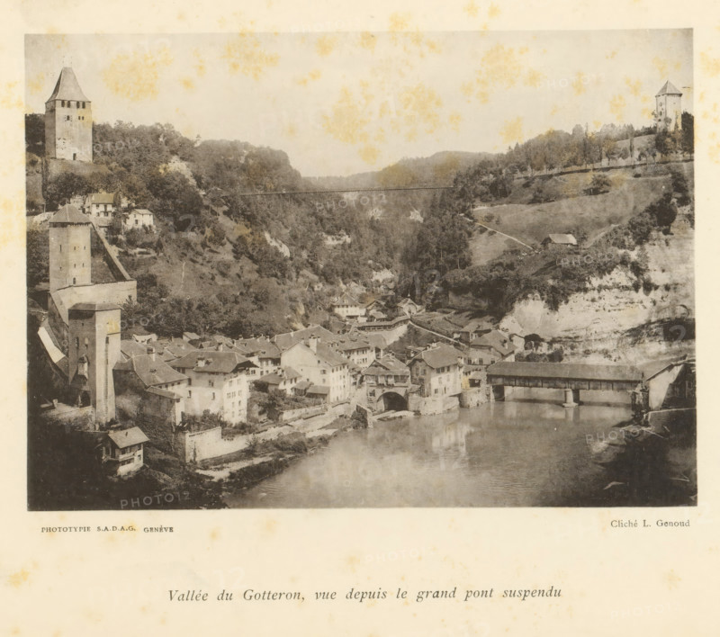 Vallée du Gotteron, vue depuis le grand pont suspendu, View of the Vallée du Gottéron from the grand pont suspendu in Fribourg, page, photomechanical print, maker: L. Genoud, Fribourg, maker: SADAG, Geneva, 1897, paper, collotype, photoprint, height 113 mm x width 157 mm, city-view, and landscape with man-made constructions, dale, valley, A sepia-toned photograph showing a valley with a river in the foreground and a clustered village of stone buildings with pitched roofs along its banks, a covered wooden bridge crossing the river, several tall stone towers on the hillsides, terraced slopes and trees behind the village, a narrow suspension bridge spanning the gorge in the distance, and a hillside road leading to a small isolated building.
