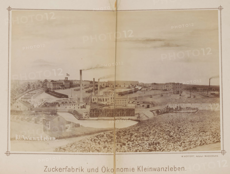 View of a sugar factory in Klein Wanzleben, Zuckerfabrik und Ökonomie Kleinwanzleben, photograph, page, photographer: Wilhelm Höffert, Klein Wanzleben, c. 1884 - in or before 1894, Saxony-Anhalt, paper, albumen print, height 129 mm x width 189 mm, Photo on frontispiece., factory in landscape, sugar, façade (of house or building), A wide sepia-toned panoramic image of an industrial complex set in a rural landscape, featuring multiple long low buildings and larger multi-story factory structures, tall smokestacks emitting plumes of smoke, rows of smaller dwellings along a foreground road, fenced yards and open storage areas, a body of water with adjacent scaffolding or framed construction, terraces of cultivated fields and dense vegetation on rolling ground, scattered trees and distant low hills under a pale sky with faint cloud formations, and a decorative border framing the scene.