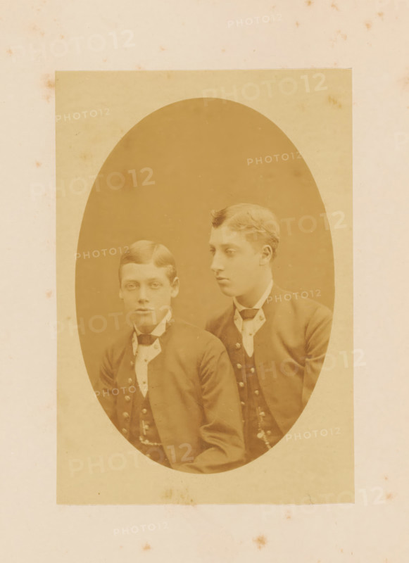 Portrait of Prince George of Wales and Prince Albert Victor of Wales, photograph, page, 1881, photographer, paper, albumen print, height 145 mm x width 99 mm, Person, George V (King of the United Kingdom), Albert Victor (Prince of the United Kingdom), prince, crown prince, A sepia-toned oval portrait mounted on a rectangular piece of paper shows two young men seated closely together, both wearing dark jackets with rows of buttons and white high-collared shirts with cravats; the man on the left faces forward with a neutral expression and neatly parted hair, while the man on the right is turned slightly toward the left with a similar neutral expression and waved hair.
