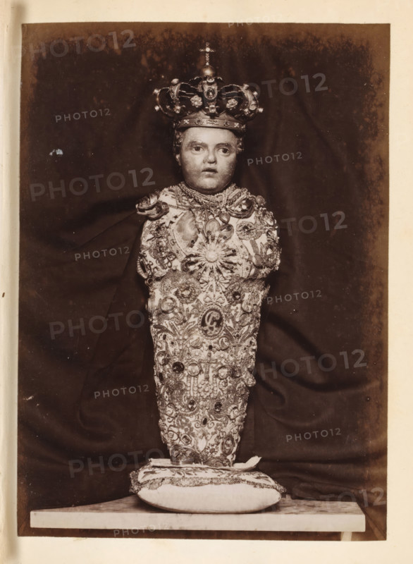 Wax figure of a baby with a crown and decorated with gemstones, photograph, page, c. 1850 - in or before 1860, photographer, paper, albumen print, height 135 mm x width 97 mm, piece of sculpture, reproduction of a piece of sculpture, A vertically oriented sepia photograph showing a single bustlike figure centered against a dark draped background, wearing an ornate crown with rounded arches and decorative elements, a lifelike face with neutral expression, and a heavily embellished garment covered in intricate embroidery, jewels, and medallionlike ornaments; the figure rests on a decorative cushion placed on a flat surface, and the image is bordered by a lighter margin.