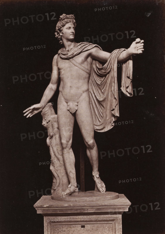 Apollo Belvedere in the Vatican Museums, photograph, page, Vatican Museums, c. 1850 - in or before 1860, photographer, Vatican City, paper, albumen print, height 134 mm x width 96 mm, (story of) Apollo (Phoebus), A photograph of a marble statue of a nude male figure standing on a rectangular plinth, shown against a dark background; the figure has curly hair and a composed facial expression, a draped garment over one shoulder and arm that falls in folds, an outstretched arm with an open hand, and the opposite arm relaxed with fingers extended; a leafy covering adorns the groin area, one leg is slightly forward with the foot wearing a detailed sandal, and a carved support column with decorative elements stands beside the figure, resting on the round base atop the plinth.