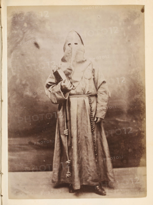 Portrait of someone in a monk's habit and with a hood over their head, photograph, page, Italy, c. 1850 - in or before 1860, photographer, paper, albumen print, height 136 mm x width 98 mm, monks and nuns, A sepia-toned photograph of a person standing against a plain backdrop wearing a full-length hooded robe with a pointed hood that covers the face except for two eye openings, a belt tied at the waist with a cord and hanging tassels, a strand of beads hanging from the waist, and a circular patch on the chest; the person holds a rectangular object in one hand and a staff or pole in the other, and wears dark shoes visible beneath the hem of the robe.