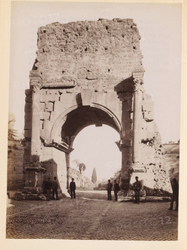 View of an ancient city gate of Rome, photograph, page, Rome, c. 1850 - in or before 1860, photographer, paper, albumen print, height 134 mm x width 97 mm, city-gate, A large ancient stone archway with weathered blocks and columns fills the frame, showing a rounded arch opening and a rough textured upper wall. The arch rests on thick supports with visible carvings and broken edges, and fragments of stone and rubble sit at the base. A cobbled street stretches beneath the arch, leading to a distant tree visible through the opening. Several men in period clothing and hats stand and pose near the arch and along the sides, some leaning against the stone and others standing on the pavement. The lighting casts shadows across the arch and the street, and portions of adjoining masonry walls are visible on either side of the structure.
