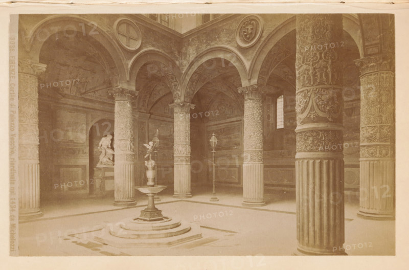 Firenze. Cortile di Palazzo Vecchio, riordinato nel 1434 da Michelozzi, Courtyard of the Palazzo Vecchio in Florence, photograph, page, number: '3036.', photographer: Edizioni Brogi (possibly), Palazzo Vecchio, c. 1878 - in or before 1888, Florence, Tuscany, paper, albumen print, height 94 mm x width 147 mm, inner court of palace or 'hôtel', A vaulted interior with multiple decorated columns supporting rounded arches, a central raised fountain with a pedestal and basin, ornate carved column capitals and patterned column shafts, a sculpted figure in an alcove to the left, two tall lamppost-like fixtures near the fountain, wall panels and painted or carved arch surfaces, a small window letting light through on the right, and a patterned floor with stepped circular base beneath the fountain.