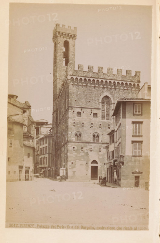 Firenze. Palazzo del Podestà o del Bargello, construzione che risale a 1250, Exterior of the Palazzo del Podestà in Florence, photograph, page, number: '3042.', photographer: Edizioni Brogi (possibly), Florence, c. 1878 - in or before 1888, Tuscany, paper, albumen print, height 145 mm x width 93 mm, façade (of house or building), hôtel, i.e. small palace in city, Palazzo del Podestà (Florence), A large stone building with a tall crenellated tower rising above a rectangular facade occupies the center of the image, its masonry showing varied stone blocks and narrow arched windows with decorative tracery, while the upper section features a row of small arches beneath battlements; adjacent to the right, a multi-story building with shuttered windows and a sloped roof stands at the street edge, and to the left a row of narrower buildings with tiled roofs and facades of different heights lines a narrow street; the foreground shows an open paved area with a few small figures and a cart near th...