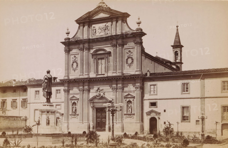 Firenze. Facciata della chiesa di S. Marco, opera dei 1780 di Giovacchino Pronti, Exterior of the Basilica di San Marco in Florence, photograph, page, photographer: Edizioni Brogi (possibly), Florence, c. 1878 - in or before 1888, Tuscany, paper, albumen print, height 92 mm x width 148 mm, church (exterior), facade (of house or building), San Marco, A sepia-toned photograph of a large ornate church façade with decorative columns, sculpted reliefs, and statues set in niches, topped by a triangular pediment and ornamental urns, flanked on the right by a bell tower with a pointed spire; in front of the church a tall statue of a robed figure stands on a multi-tiered pedestal within a fenced garden area containing small plants and lampposts, and adjacent buildings with shuttered windows form the background.