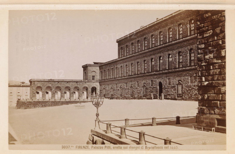 Firenze. Palazzo Pitti, eretto sui disegni di Brunellesco nel 1440, Facade of the Palazzo Pitti in Florence, photograph, page, number: '3037bis.', photographer: Edizioni Brogi (possibly), Florence, c. 1878 - in or before 1888, Tuscany, paper, albumen print, height 93 mm x width 143 mm, façade (of house or building), hôtel, ie small palace in city, Palazzo Pitti, A wide courtyard bordered by a large stone palace with rusticated masonry and rows of arched windows across multiple stories, an arched colonnade extending from one side, a raised stone terrace with balustrade and low posts in the foreground, an ornate street lamp near the terrace edge, shadowed doorways and window grilles on the palace facade, and a textured stone wall framing the right edge of the scene.