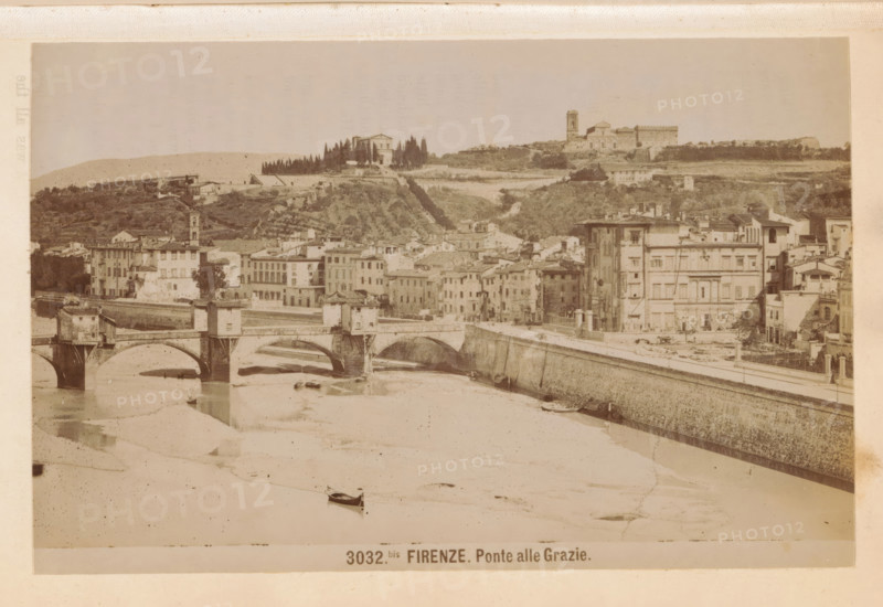 Firenze. Ponte alle Grazie, View of the Ponte alle Grazie in Florence, photograph, page, number: '3032bis.', photographer: Edizioni Brogi (possibly), Florence, c. 1878 - in or before 1888, Tuscany, paper, albumen print, height 95 mm x width 145 mm, bridge in city across river, canal, Ponte alle Grazie, A wide sepia-toned cityscape showing a river with arched stone bridges crossing it, small boats on the water, and a long stone embankment along the riverbank. Along the embankment a row of multi-story buildings with varied facades, windows, and rooftops rise from the street, with one taller building near the center-right that has decorative vertical features. Behind the riverside buildings a hillside with terraced greenery and scattered houses leads up to a ridge topped by a cluster of trees and larger structures, including a building with a tower and another with a rectangular, castle-like appearance. The sky above is pale and mostly featureless.