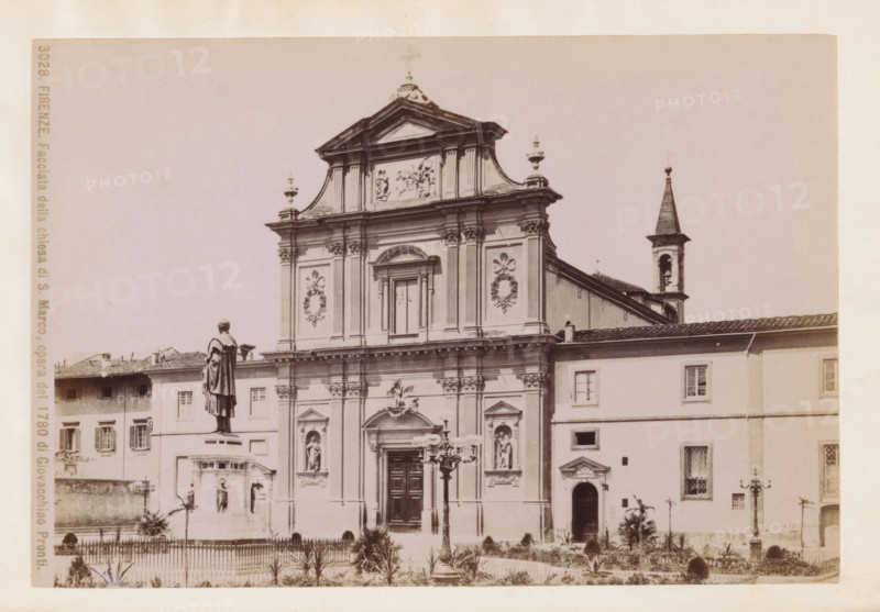 Firenze. Facciata della chiesa di S. Marco, opera del 1780 di Giovacchino Pronti, Facade of the San Marco Monastery in Florence, photograph, page, number: '3028.', photographer: Fratelli Alinari (possibly), Florence, c. 1877 - in or before 1887, Tuscany, paper, albumen print, height 98 mm x width 145 mm, Photo on frontispiece., facade (of house or building), San Marco, A sepia-toned photograph of a classical church façade with ornate architectural details including columns, pilasters, sculpted reliefs, and decorative wreath motifs, a central arched doorway with carved panels, niche statues set into the façade, and a pedimented upper section topped with finials; a bell tower with a pointed roof appears to the side; in front of the church a raised statue on a pedestal overlooks a fenced garden area with small plants and pathways, and a decorative streetlamp stands near the entrance; adjacent buildings with shuttered windows flank the church.