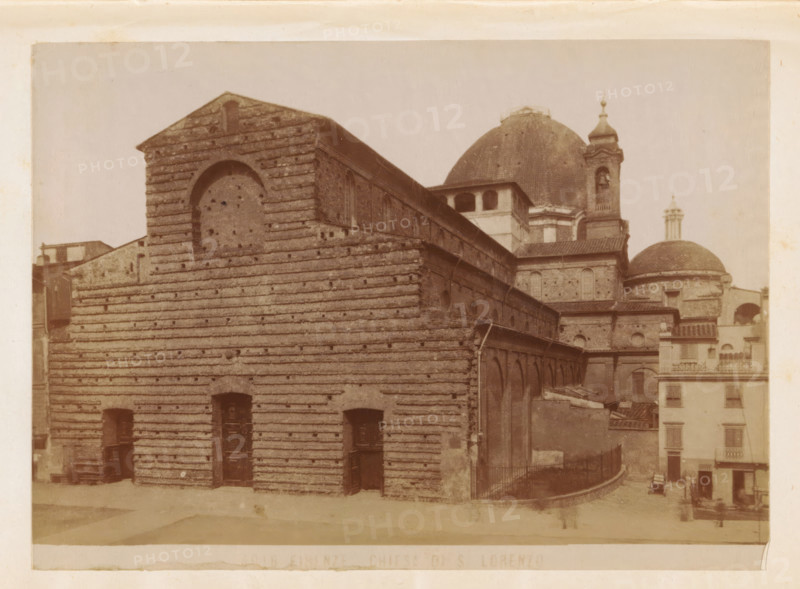 Firenze. Chiesa di S. Lorenzo, Exterior of the Basilica of St. Laurence in Florence, photograph, page, number: '4016', Florence, c. 1880 - in or before 1890, photographer, Tuscany, paper, albumen print, height 100 mm x width 137 mm, church (exterior), facade (of house or building), St. Lawrence Basilica, A sepia-toned photograph of a large stone building with horizontal banding and a triangular gable, showing arched and rectangular openings and wooden doors at ground level, with a long side façade featuring a row of small arched windows and an upper level with larger openings; behind and to the right there are domed roofs, one large dome with a smaller dome and a bell tower topped by a decorative finial, and adjacent multi-story buildings with balconies and shuttered windows; the foreground shows a paved area and a curved railing leading toward the rear, with faint human figures and a horse-drawn cart near the right edge.