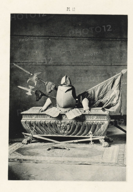 Sarcophagus with armor and weapons, page, photomechanical print, number: 'Pl 42', Barcelona (city), c. 1880 - in or before 1881, Catalonia, paper, collotype, photoprint, height 147 mm x width 103 mm, weapons, sarcophagus, harness, A photographic print shows a decorative stone or carved base with scroll and floral motifs resting on lion paw feet, atop which sits a suit of armor including a breastplate and a helmet with a brim. Pole weapons with ornate heads and tassels are arranged behind and across the armor, and a draped fabric flag with visible folds is positioned to one side. In front of the base, crossed swords rest on the floor over a patterned rug with a central medallion design.