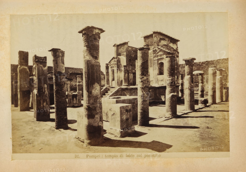 Pompeii: tempio di Iside col peristilio, Ruin of the Tempio di Iside in Pompeii, photograph, page, number: '32.', c. 1878 - in or before 1888, photographer, paper, albumen print, height 103 mm x width 153 mm, ruin of a building, architecture, temple, shrine, Roman religion, Tempio di Iside, A sepia photograph shows a sunlit archaeological site with rows of weathered stone columns and rectangular stone blocks arranged on a sandy ground, some columns topped with rough capitals and others with flat fragments, casting long shadows; in the background there are ruined stone walls and a partially intact façade featuring an arched opening and decorative pediment, with worn brick and plaster textures visible across the structures.