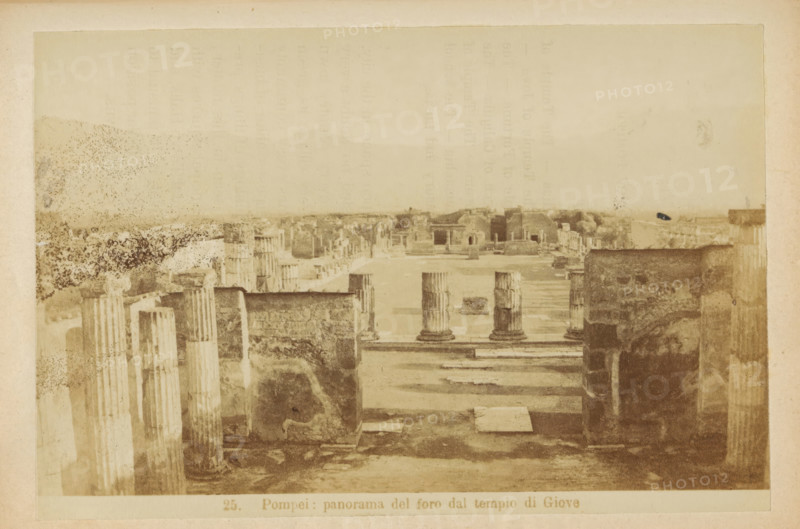 View of the Foro Civile at Pompeii, Pompeii: panorama of the foro dal tempio di Giove, photograph, page, c. 1878 - in or before 1888, photographer, paper, albumen print, height 104 mm x width 153 mm, market, landscape with ruins, Foro Civile, A sepia-toned photograph shows an ancient open area with rows of weathered stone columns and ruins. In the foreground there are tall fluted columns and rectangular stone bases, some broken and partly eroded. A wide paved walkway leads toward a central open plaza where a few short column stumps stand on square platforms. Low stone walls and fragmentary structures flank the plaza, and beyond them a distant horizontal line of additional ruined buildings and foundations is visible under a pale sky. The image has uneven staining and spotting across the surface and a faded border around the edges.