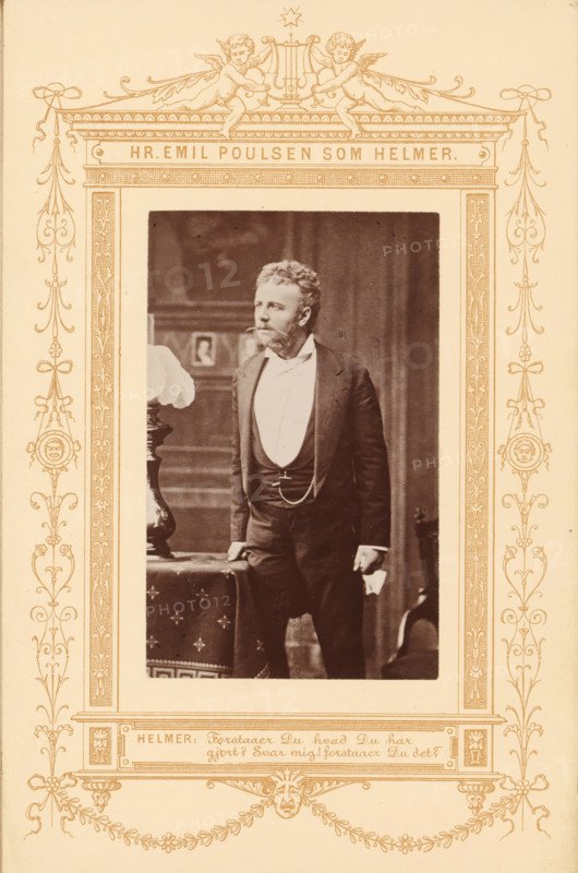 Hr. Emile Poulsen som Helmer, Portrait of the actor Emil Poulsen as Helmer in A Doll's House, photograph, page, inscription: 'Helmer:, Du det?', photographer: JA Schulz, printer (person): Edvin Jantzen, publisher: Ernst Bojesen, c. 1870 - in or before 1880, JA Schulz, Edvin Jantzen, Ernst Bojesen, photographer, printer (person), publisher, paper, albumen print, albumendruk, height 94 mm x width 60 mm, Person, Emil Poulsen, portrait of actor, actress, A sepia photograph shows a man standing indoors, dressed in a dark suit with a waistcoat and a white shirt, his hand resting on a table draped with a patterned cloth and the other hand holding a folded item; he has a beard and curly hair and looks slightly to his left. The framed portrait is set within an ornate decorative border featuring symmetrical floral and scroll motifs, medallion faces, ribbon-like elements, and two cherubs at the top holding a lyre, all rendered in a single ink color against a light background. The interior backgro...