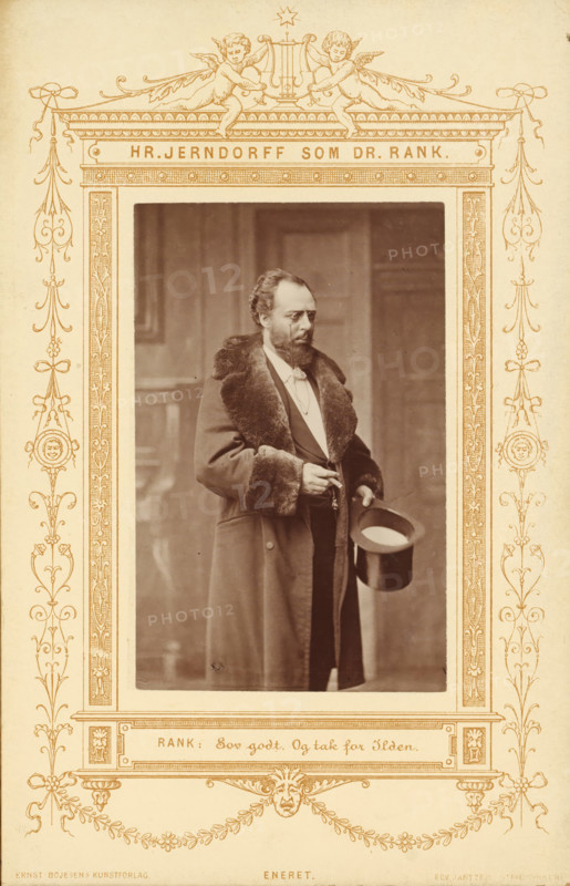 Portrait of the actor Peter Jerndorff as Doctor Rank in A Doll's House, Hr. Jerndorff sum Dr. Rank, photograph, page, inscription: 'Rank:, Ilden.', photographer: JA Schulz, printer (person): Edvin Jantzen, publisher: Ernst Bojesen, c. 1870 - in or before 1880, JA Schulz, Edvin Jantzen, Ernst Bojesen, photographer, printer (person), publisher, paper, albumen print, albumendruk, height 99 mm x width 62 mm, Person, Peter Jerndorff, portrait of actor, actress, A sepia-toned framed photograph shows a man standing indoors wearing a long coat with a fur collar and fur cuffs, a suit underneath, and a bow tie; he has a beard and mustache and is holding a top hat in one hand while the other hand holds a small object near his waist; the decorative frame around the photograph features symmetrical ornamental flourishes, cherub figures at the top, and intricate border patterns on a light background.