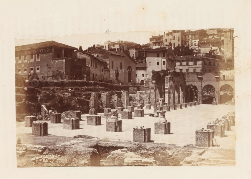 View of the ruins of the Basilica Julia on the Roman Forum in Rome, photograph, page, Roman Forum, c. 1863 - in or before 1873, photographer, Rome, Forum Romanum, paper, albumen print, height 92 mm x width 150 mm, ruin of a building, architecture, Basilica Julia, A sepia-toned photograph showing a scene of ruins and old buildings with an open foreground filled with rectangular stone blocks arranged in rows, some with debris on top, and a few shallow excavated areas. Behind the blocks a raised embankment leads to a row of multi-story buildings with tiled roofs, shuttered windows, and a central structure with a tall arched doorway. To the right a long arcade of arches and columns connects to more masonry and a ladder leaning against one arch. Scattered stones and rough terrain occupy the lower part of the image, and the sky above is pale and untextured.