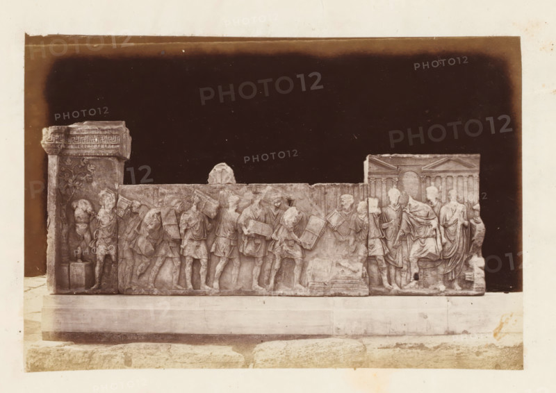 Relief on the Roman Forum in Rome, depicting Emperor Trajan destroying the tax documents, photograph, page, Roman Forum, c. 1863 - in or before 1873, photographer, Rome, paper, albumen print, height 100 mm x width 148 mm, official acts of rulers, eg royal acts (ruler giving audience, granting mercy, etc., ruler and his subjects), sculpture (relief, sculpture), taxes and financial administration, A sepia-toned photograph shows a long stone relief mounted on a low plinth against a dark background, depicting a procession of carved human figures in classical dress. The relief includes a column fragment at one end with decorative carving and a vertical vine motif, a row of standing and walking figures across the central section many holding rectangular objects, and a group of robed figures near the other end where architectural elements with columns and a pediment are visible. The carved figures vary in posture and gesture, some looking forward, others turned toward each other, and the surf...