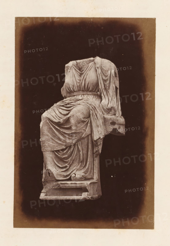 Sculpture on the Palatine Hill, depicting an enthroned Magna Mater, photograph, page, Rome, c. 1863 - in or before 1873, photographer, paper, albumen print, height 150 mm x width 100 mm, sculpture, sitting figure - AA - female human figure, A worn marble statue of a seated draped figure is centered against a dark rectangular background with a lighter border; the figure lacks a head and shows detailed folds of cloth gathered at the waist and cascading over the legs and seat, with one visible foot wearing a sandal resting on a block and parts of the statue showing chips and weathering.