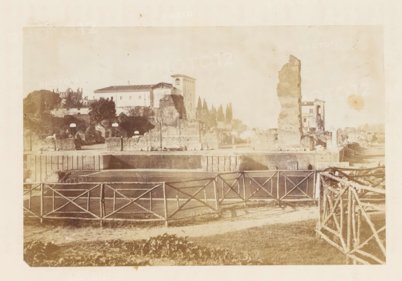 View of ruins (presumably) at Rome, photograph, page, Rome, c. 1863 - in or before 1873, photographer, paper, albumen print, height 100 mm x width 140 mm, landscape with ruins, A sepia-toned view of an open area with a wooden lattice fence in the foreground and a pathway or road behind it, leading toward ruins and stone structures across the middle ground; there are tall broken stone walls and fragments, a rectangular building with a pitched roof and a taller tower-like building behind it, and a taller irregular stone column standing near a partially intact building with arched openings; scattered trees and cypress-like vertical trees rise behind the ruins, and low vegetation and grass appear in the front area, with an overall faded, aged appearance.