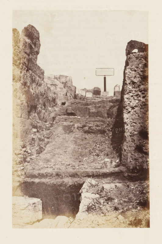 Ruin on the Palatine Hill in Rome, photograph, page, Palatine Hill, c. 1863 - in or before 1873, photographer, Rome, paper, albumen print, height 153 mm x width 96 mm, ruin of a building, architecture, A sepia-toned photograph showing a rough, uneven pathway carved between tall, rocky walls that rise on both sides, with the left wall appearing jagged and the right wall textured and crumbling; the ground is strewn with small stones and patches of sparse vegetation, and a stepped ledge is visible near the foreground; in the distance a freestanding rectangular sign mounted on a post stands on a small base among more rocky outcrops.