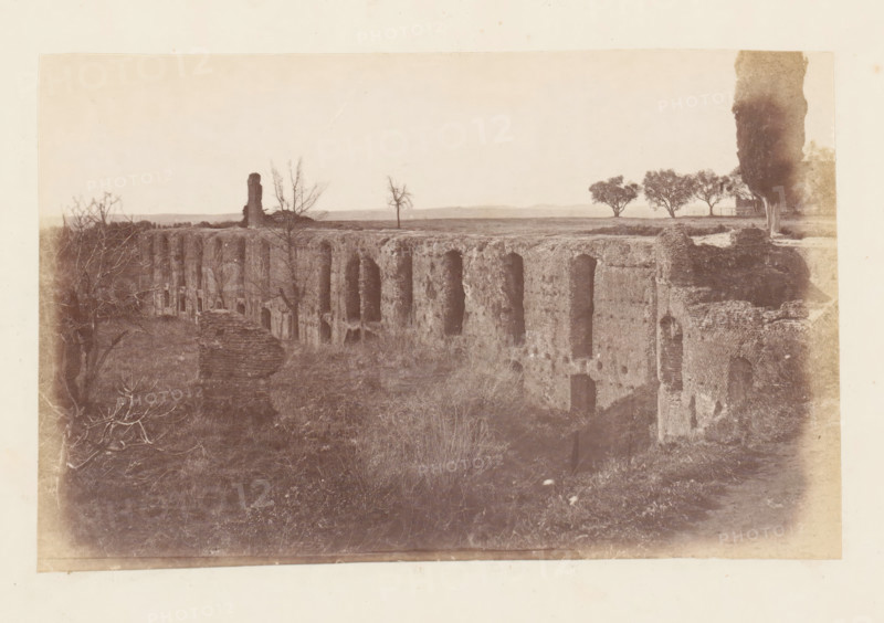 View of the ruins of the Villa Adriana at Tivoli, photograph, page, Tivoli, c. 1863 - in or before 1873, photographer, paper, albumen print, height 95 mm x width 153 mm, villa, ruin of a building, architecture, Villa Adriana, A wide sepia-toned photograph showing a long, weathered stone structure with a series of tall arch-shaped openings along its vertical face, set in an open grassy area. In front of the structure there is overgrown vegetation and low shrubs. Leafless tree branches appear on the left foreground, and a lone bare tree stands on the flat top of the structure. On the right side a tall, narrow tree rises above the ground near a small fence and a cluster of fuller trees further back. The horizon is low and distant, with a pale sky occupying most of the upper portion of the image.