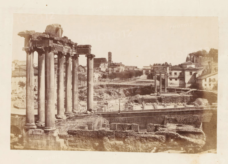 View of the Roman Forum in Rome, with the ruins of the Temple of Saturn in the foreground, photograph, page, Roman Forum, c. 1863 - in or before 1873, photographer, Rome, paper, albumen print, height 100 mm x width 153 mm, landscape with ruins, temple, shrine, Roman religion, Temple of Saturn, A sepia-toned photograph showing tall classical columns with ornate capitals and a partially ruined entablature in the foreground, standing on a stone base above excavated ruins and stone foundations. In the middle distance there are additional isolated columns and fragments scattered among open trenches and low walls. A paved roadway or promenade lined with trees runs behind the ruins, with a mix of masonry buildings, a tower, and tiled-roof structures along its edge. The sky appears pale and empty above the scene, and the image has a wide margin of lighter paper around the printed area.