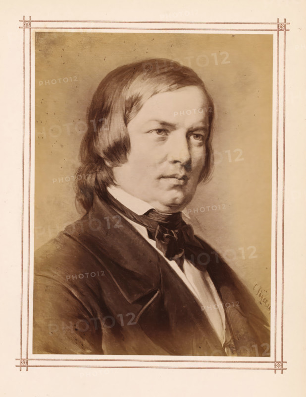 Photo reproduction of a portrait of Robert Schumann by Carl Jaeger, photograph, reproduction, page, name: 'C. Jäger 18...', after painting by Carl Jaeger, c. 1865 - c. 1875, photographer, paper, albumen print, height 144 mm x width 106 mm, Person, Robert Schumann, composer, A sepia-toned portrait of a man shown from the chest up, with medium-length straight hair swept to the sides, a serious facial expression, and visible eyes looking slightly off center, wearing a dark jacket over a white shirt and a dark tied scarf at the collar, set against a plain shaded background and framed by a decorative double-lined border.