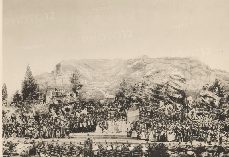 Festakt. Schlussbild, View of the final performance of the Calven-Feier in Chur, page, photomechanical print, number: 'V.', photographer: Lienhard & Salzborn, Chur, c. 1898 - in or before 1899, paper, collotype, photoprint, height 180 mm x width 254 mm, group of actors, troupe, actors on the stage, mountains, A wide sepia-toned scene shows a large outdoor gathering set against a backdrop of rocky mountains and trees, with a multitude of people assembled on tiered stone steps and rocky outcrops. Many individuals wear varied historical costumes and garments, including long dresses, cloaks, and headwear, and several people hold large flags and banners. In the foreground a group of musicians with instruments sit behind a low barrier, and in front of them performers stand on a raised platform where a few figures with distinctive robes and shields are positioned. To the sides and behind the platform the crowd rises densely, interspersed with tall evergreen trees, rocky formations, and what a...