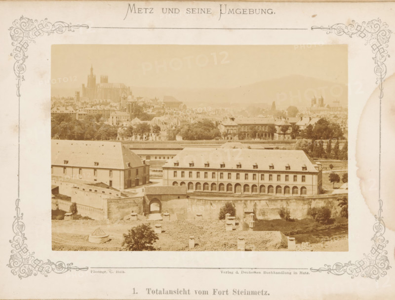 View of Metz, Total view of Fort Steinmetz, Metz and Seine Umgebung, photograph, page, number: '1.', photographer: C. Holt, Metz, publisher: Verlag der Deutschen Buchhandlung, c. 1865 - in or before 1875, paper, albumen print, albumendruk, height 99 mm x width 141 mm, Photo opposite title page., city-view, and landscape with man-made constructions, A sepia-toned panoramic view framed by decorative borders shows a townscape with a large multi-story building in the foreground featuring a row of arched openings on the ground level and dormer windows on a sloped roof, set behind a fortified wall with an arched gateway. Behind this complex are rows of trees and additional buildings with varied roofs and architectural shapes, and further back a dense cluster of houses and taller structures including a cathedral-like building with towers and steep vertical elements. Hills rise faintly in the distant background under a pale sky, and the entire scene has an aged, antique photograph appearance.
