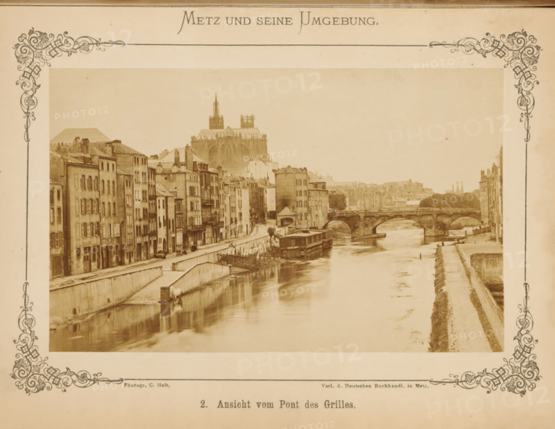 Ansicht vom Pont des Grilles, View of Metz and the Moselle, Metz und seine Umgebung, photograph, page, number: '2.', photographer: C. Holt, Metz, publisher: Verlag der Deutschen Buchhandlung, c. 1865 - in or before 1875, paper, albumen print, albumendruk, height 96 mm x width 155 mm, city-view, and landscape with man-made constructions, bridge in city across river, canal, A sepia-toned photographic scene framed by decorative borders shows a river flowing between stone embankments with rows of multi-story buildings lining one side and a walkway along the opposite side, a stone arched bridge spans the river in the middle distance, boats and a riverside structure are moored near the bank, and a large Gothic cathedral with towers rises above the buildings in the background.