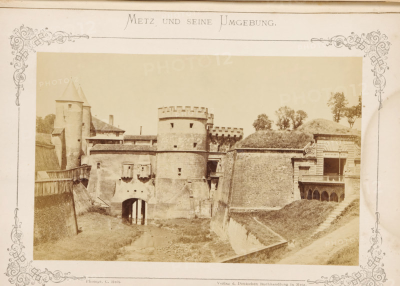 Das Deutsche Thor, View of the Porte des Allemands in Metz, Metz und seine Umgebung, photograph, page, number: '5.', photographer: C. Holt, Metz, publisher: Verlag der Deutschen Buchhandlung, c. 1865 - in or before 1875, paper, albumen print, albumendruk, height 99 mm x width 155 mm, city-gate, Porte des Allemands, A sepia-toned photograph shows a fortified stone structure with rounded towers and battlements, connected by thick walls and a bridge-like entrance over a narrow water channel, flanked by sloped earthworks and grassy embankments. The left side features tall conical-roofed towers and attached buildings with tiled roofs, while the central section displays a cylindrical keep with small window openings and crenellations. The right side includes a carved stone façade with an open balcony above arched openings, and a grassy mound with scattered trees rising behind the fortifications. Decorative ornate borders frame the photograph on all sides.
