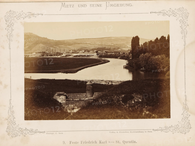Feste Friedrich Karl - St. Quentin, View of the Feste Prinz Friedrich-Karl in Metz, Metz and seine Umgebung, photograph, page, number: '9.', photographer: C. Holt, Metz, publisher: Verlag der Deutschen Buchhandlung, c. 1865 - in or before 1875, paper, albumen print, albumendruk, height 96 mm x width 143 mm, defense of fortification or city, A sepia-toned landscape photograph mounted on a decorative bordered page showing a curving river flanked by grassy banks and dense stands of tall trees on one side, with a low stone fortification with a cylindrical turret in the foreground set into a sloping hillside, and fields, industrial buildings, and rolling hills visible in the distant background under a clear sky.