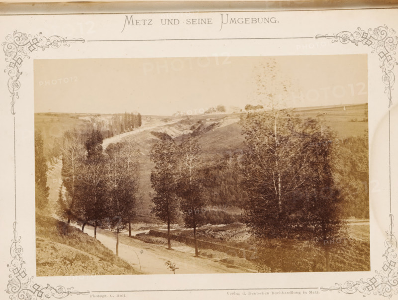 Schlucht von Gravelotte I, View of the gorge near Metz where the Battle of Gravelotte took place in 1870, Metz und seine Umgebung, photograph, page, number: '29.', photographer: C. Holt, Gravelotte, publisher: Verlag der Deutschen Buchhandlung, Metz, c. 1865 - in or before 1875, paper, albumen print, albumendruk, height 99 mm x width 153 mm, dale, valley, A sepia-toned photograph framed by an ornate decorative border shows a rural landscape with a dirt road winding beside a row of tall trees, a sloped embankment and a low stone wall alongside the road, open fields and gently rolling hills in the background, clusters of trees and a stand of taller trees further along the valley, and a sparse foreground of grasses and bushes.