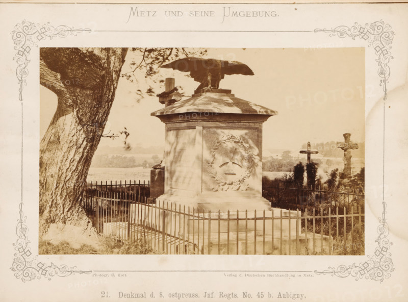Denkmal d. 8. ostpreuss. Inf. Right. No. 45 b. Aubigny, War Memorial for the 8th East Prussian Infantry Regiment No. 45 at Aubigny near Metz, Metz und seine Umgebung, photograph, page, number: '21.', photographer: C. Holt, Lorraine, publisher: Verlag der Deutschen Buchhandlung, Metz, c. 1870 - in or before 1875, paper, albumen print, albumendruk, height 98 mm x width 154 mm, monument, statue, A sepia-toned photograph shows a fenced stone monument topped by a large sculpted eagle with outstretched wings standing on a square plinth, the plinth bearing a wreath relief on one visible face. A tall tree trunk with textured bark occupies the left foreground, and a simple metal fence surrounds the monument. In the background there are landscape elements including distant trees and fields, as well as two crosses—one plain wooden cross and one carved stone cross—visible to the right of the monument. Decorative printed borders frame the photograph on the paper mount.