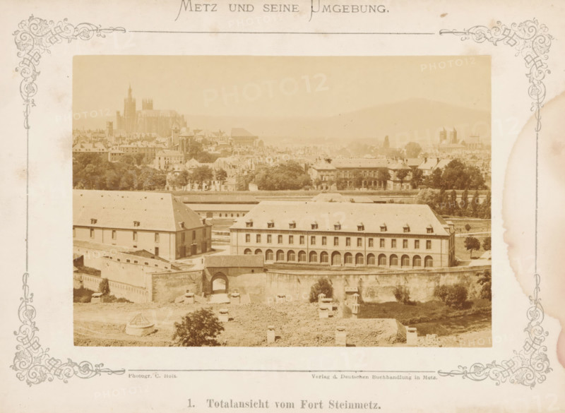 Metz und seine Umgebung, book, 1875, textile materials, pasteboard, paper, linen (material), albumen print, color on cut, height 210 mm x width 160 mm x thickness 20 mm, A sepia-toned photograph centered within a decorative border shows a panoramic urban scene with a large multi-story building in the foreground featuring a long row of arches along its ground level and steeply pitched roofs with small dormer windows, adjacent to another rectangular building with a sloped roof and small windows. A fortified wall with an arched gateway runs beneath these buildings, and open ground with scattered low structures and a small circular roofed feature lies in front. Behind the foreground complex, rows of trees and varied residential and institutional buildings fill the middle distance, with a prominent cathedral-like structure rising above the city skyline to the left and additional church towers visible toward the right, all set against hazy rolling hills in the background. The image is framed...