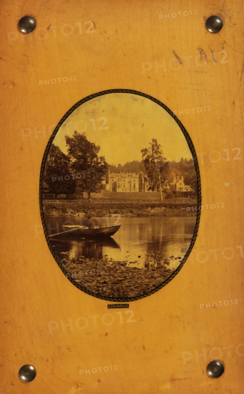 Abbotsford, View of Abbotsford House, photograph, page, c. 1858 - in or before 1868, photographer, paper, plant material, biological material, wood (plant material), albumen print, height 95 mm x width 73 mm, Photo on back cover., rowing-boat, canoe, castle, Abbotsford House, An oval sepia photograph is mounted on a worn light brown wooden board with four dark round metal studs near the corners; inside the oval, a calm river reflects a small rowboat with a seated figure holding oars, a pebbled shoreline in the foreground, a grassy riverbank with trees behind it, and a large multi-gabled building partially obscured by trees in the midground, all under a pale sky.