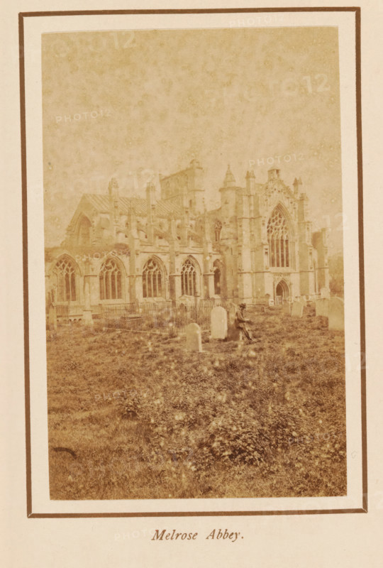 Melrose Abbey, photograph, page, photographer: George Washington Wilson, Scotland, c. 1885 - c. 1890, paper, albumen print, height 108 mm x width 68 mm, ruin of church, monastery, Melrose (Scotland), A faded sepia photograph shows the ruins of a large Gothic stone abbey with tall arched windows and detailed tracery, buttresses, and a prominent ornate façade; the building sits behind a grassy area with scattered upright tombstones and a fenced section of graves, and a solitary seated figure wearing a hat is visible among the stones near the front of the abbey.